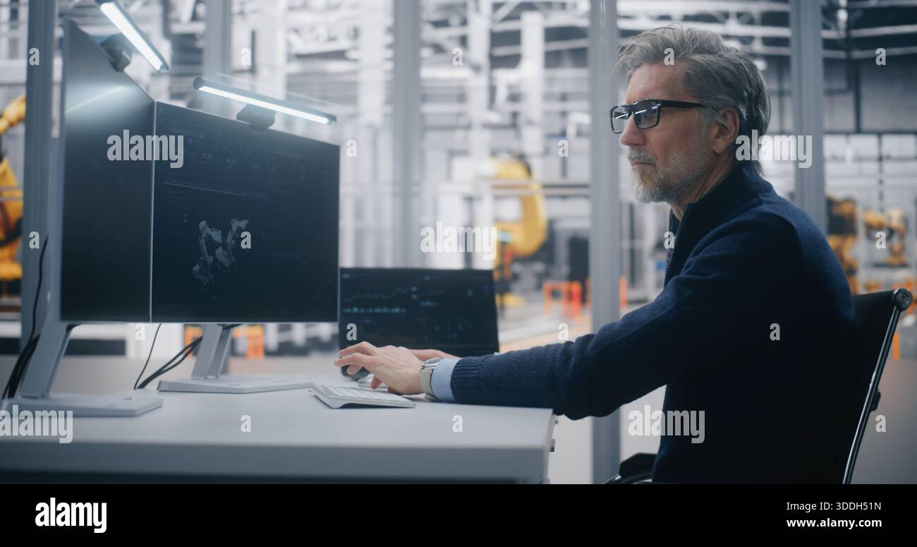 Focused Male Professional Programs Robotic Equipment While Tracking Operational Metrics on Dual Monitor Desk in Modern Factory Setting. Concept of Industrial Automation and Production Strategy. Stock Photo