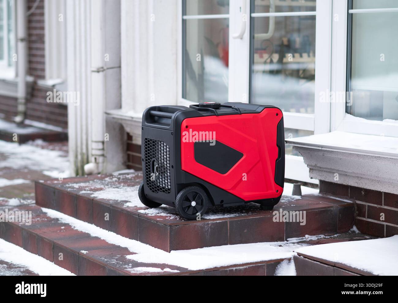 Red portable inverter generator standing on snowy stairs near a building. Backup power source, winter blackout  Stock Photo
