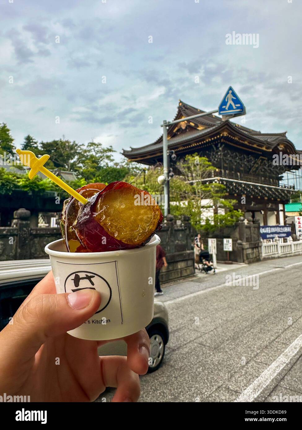 Hand holding imo kekki caramelized sweet potato snack with Narita Sōhō-ji temple gate in background, Narita, Chiba, Japan - Smartphone Captured Stock Image