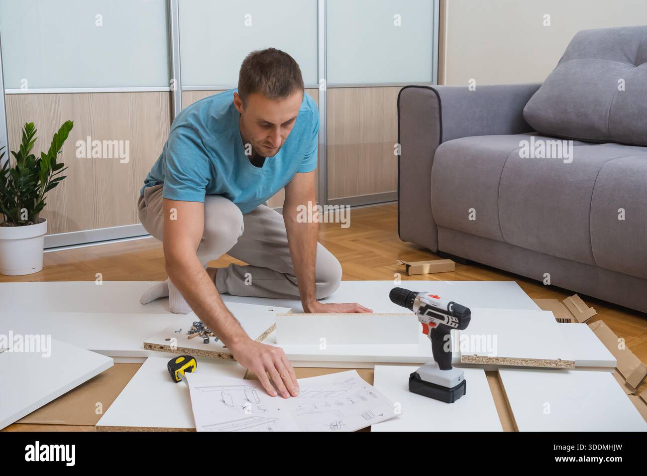 man sits on the floor reading furniture assembly instructions with tools and flat-pack parts scattered around him in a bright, modern living room Stock Photo