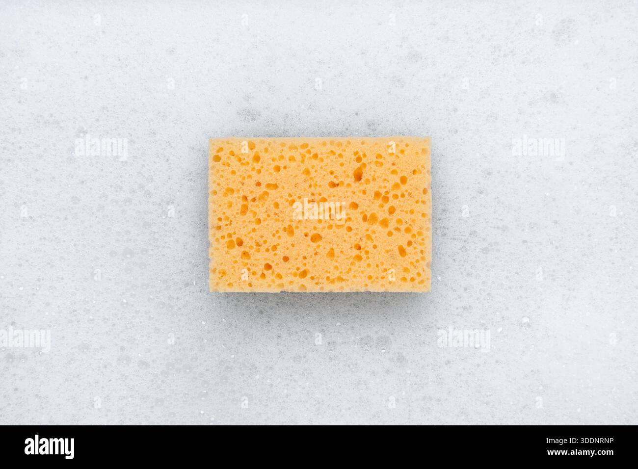 Yellow sponge on a white soapy foam background. Top view.м Stock Photo