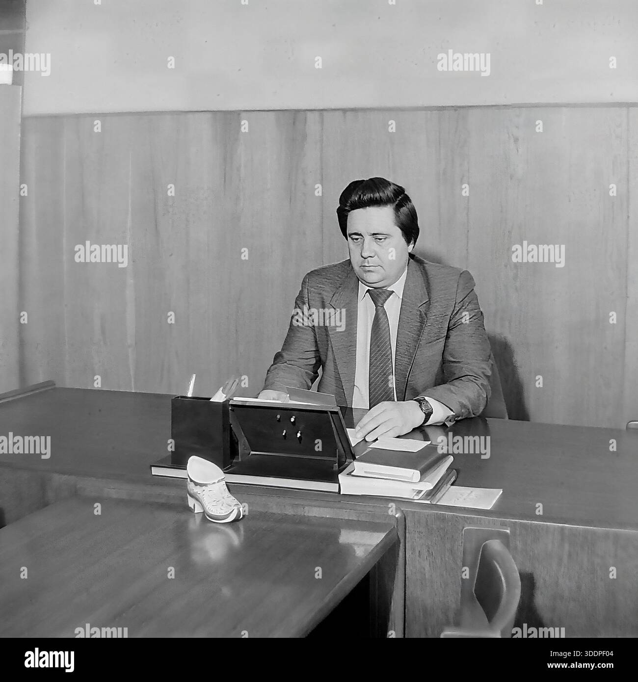 Slaviansk, Ukrainian SSR, Soviet Union, 1980s. An authentic archival working portrait of Comrade Novikov, director of the AIZ Armature and Insulator Plant, sitting at his desk in a wood-paneled office. The man is dressed in a suit and tie, focused on reading documents and handling business papers. The desk features a calendar stand, books and a decorative ceramic shoe. This vintage black and white photograph captures the daily routine of a Soviet industrial leader and the stability of the peaceful Donbas region during the USSR era. Stock Photo