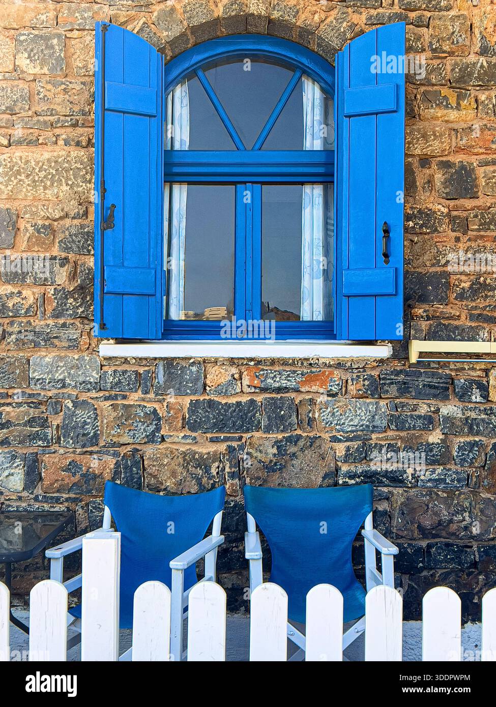 Arched window with vibrant blue shutters on a rustic stone wall, with two chairs and a white fence Stock Photo