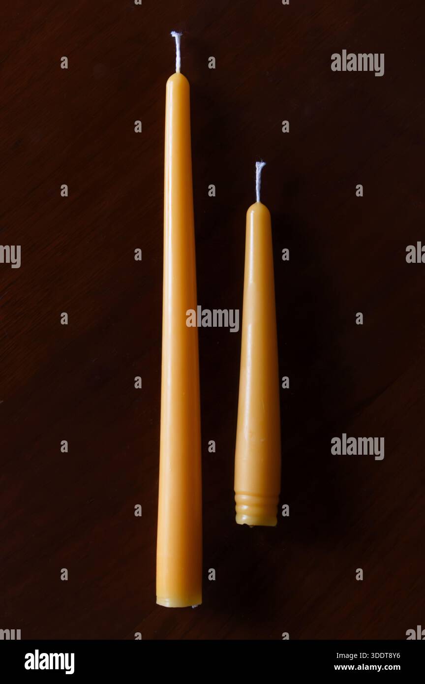 Two yellow beeswax candles of different sizes laying on a dark wood table Stock Photo