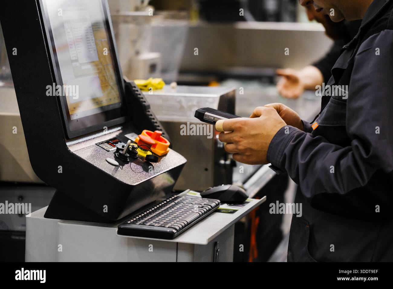 Factory technician using data collection device to log operations on CNC control console with digital display and manual controls Stock Photo