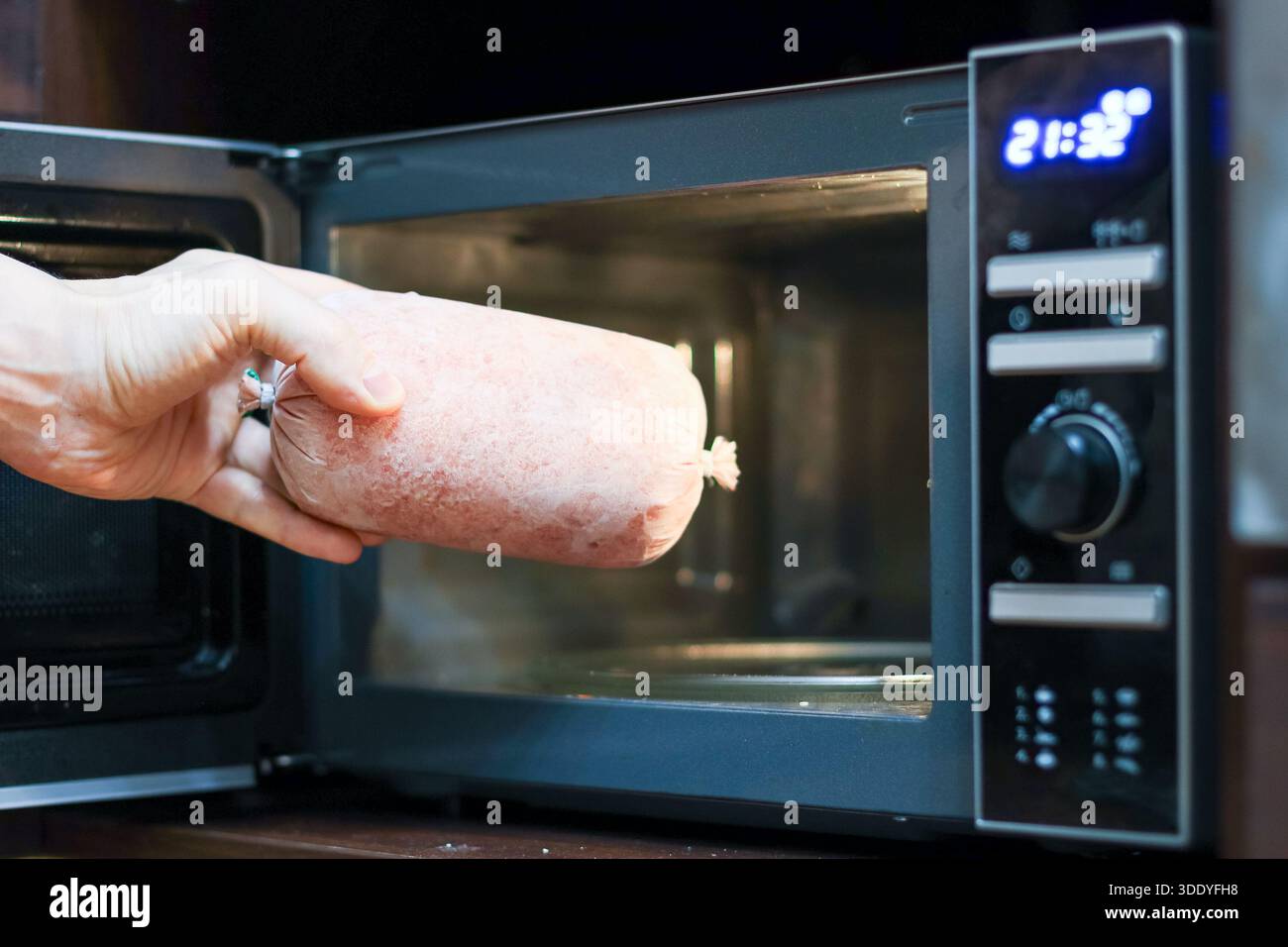Fingers grasp a iced sausage cylinder extending into the lit microwave cavity with timer display visible. Stock Photo