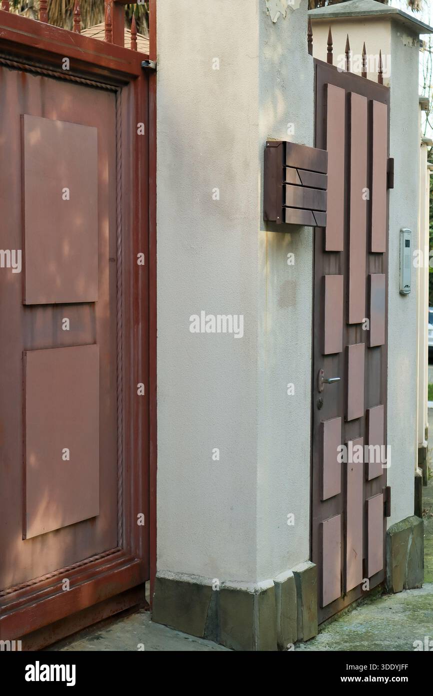 Rustic red metal pedestrian gate featuring abstract rectangular panels, mounted intercom, and spiked top on a stucco wall. Stock Photo
