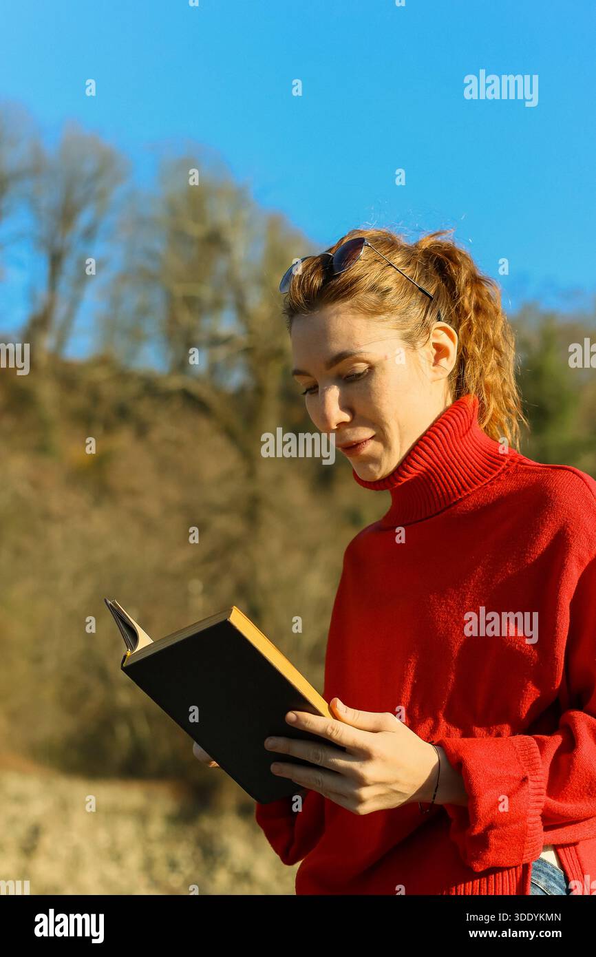 A young woman with ponytail and sunglasses on head holds an open book while standing in a natural landscape with trees and clear sky. Stock Photo