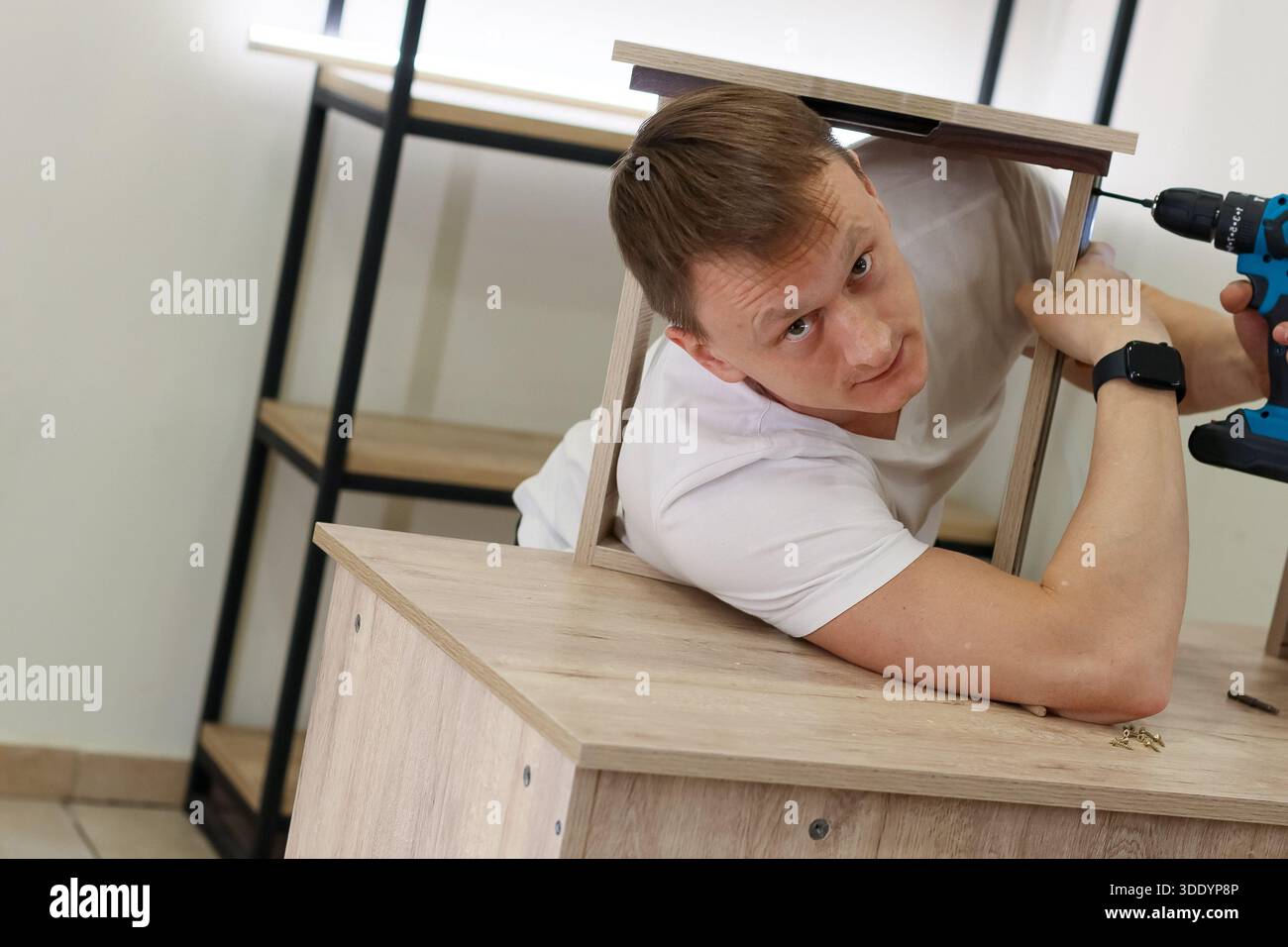 Young man in white shirt reaches under wooden frame on table with blue drill, screwing from side in challenging position indoors. Stock Photo
