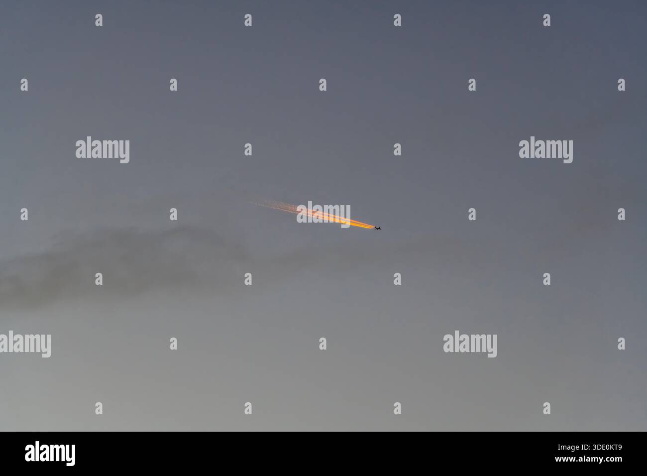 An aircraft flying high in a clear sky, with its contrail lit pink by the setting sun Stock Photo