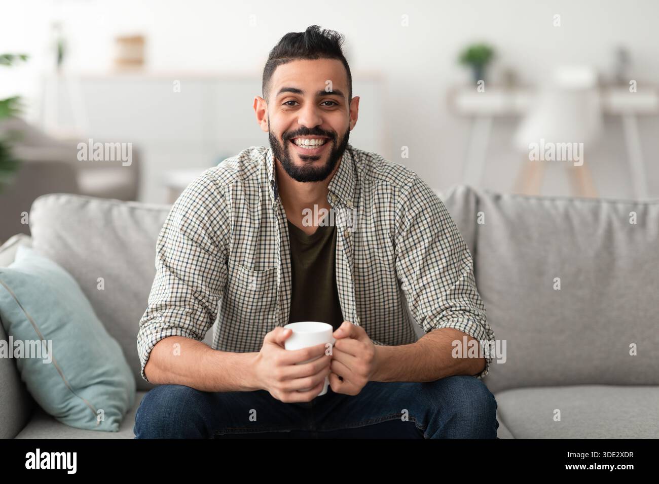 Young smiling Arab guy drinking hot aromatic coffee on sofa at home, copy space Stock Photo