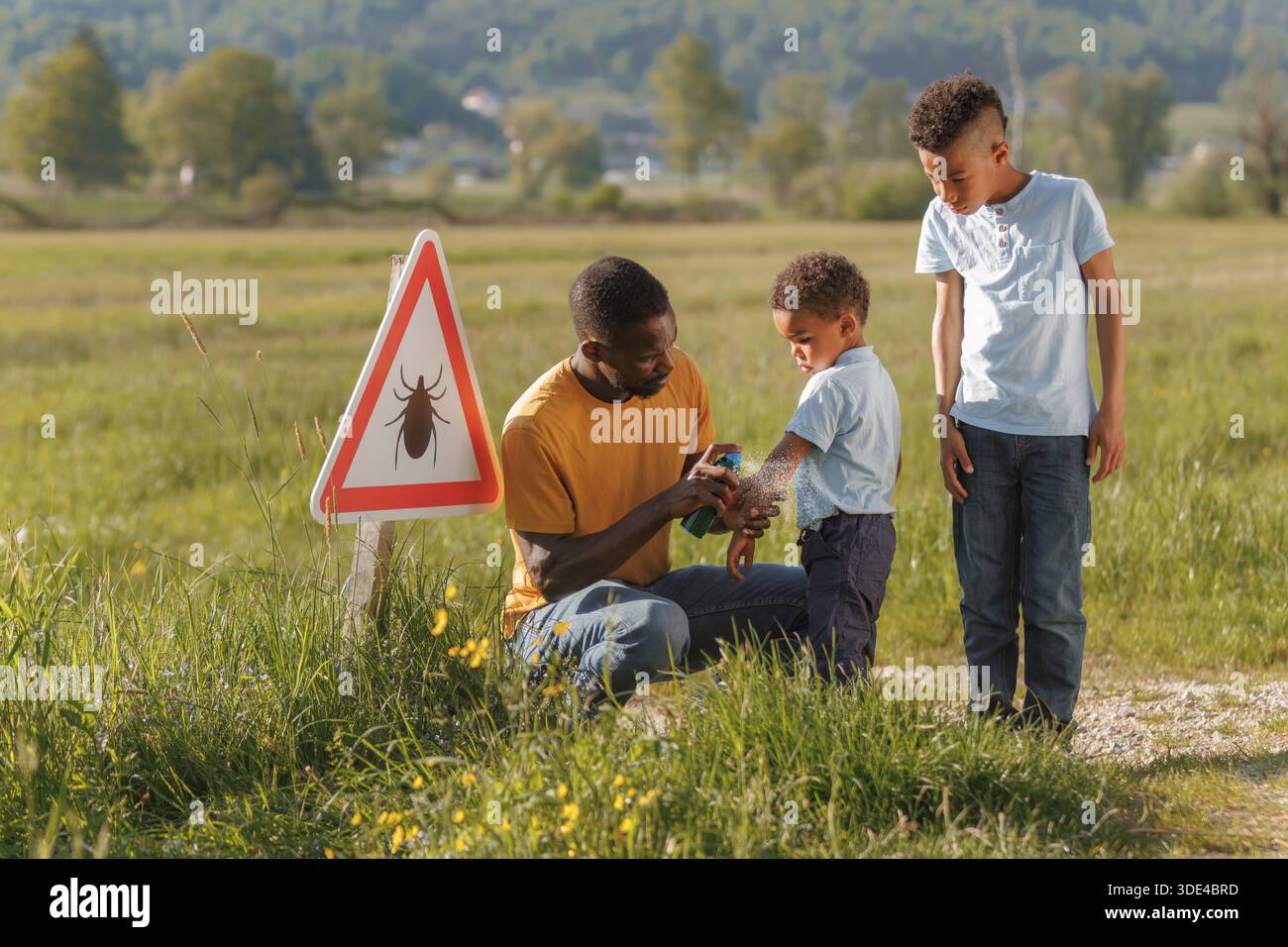 Man taking precautions against insects during a nature walk, spraying his sons near a tick awareness sign. Preventing Lyme disease infection. Stock Photo