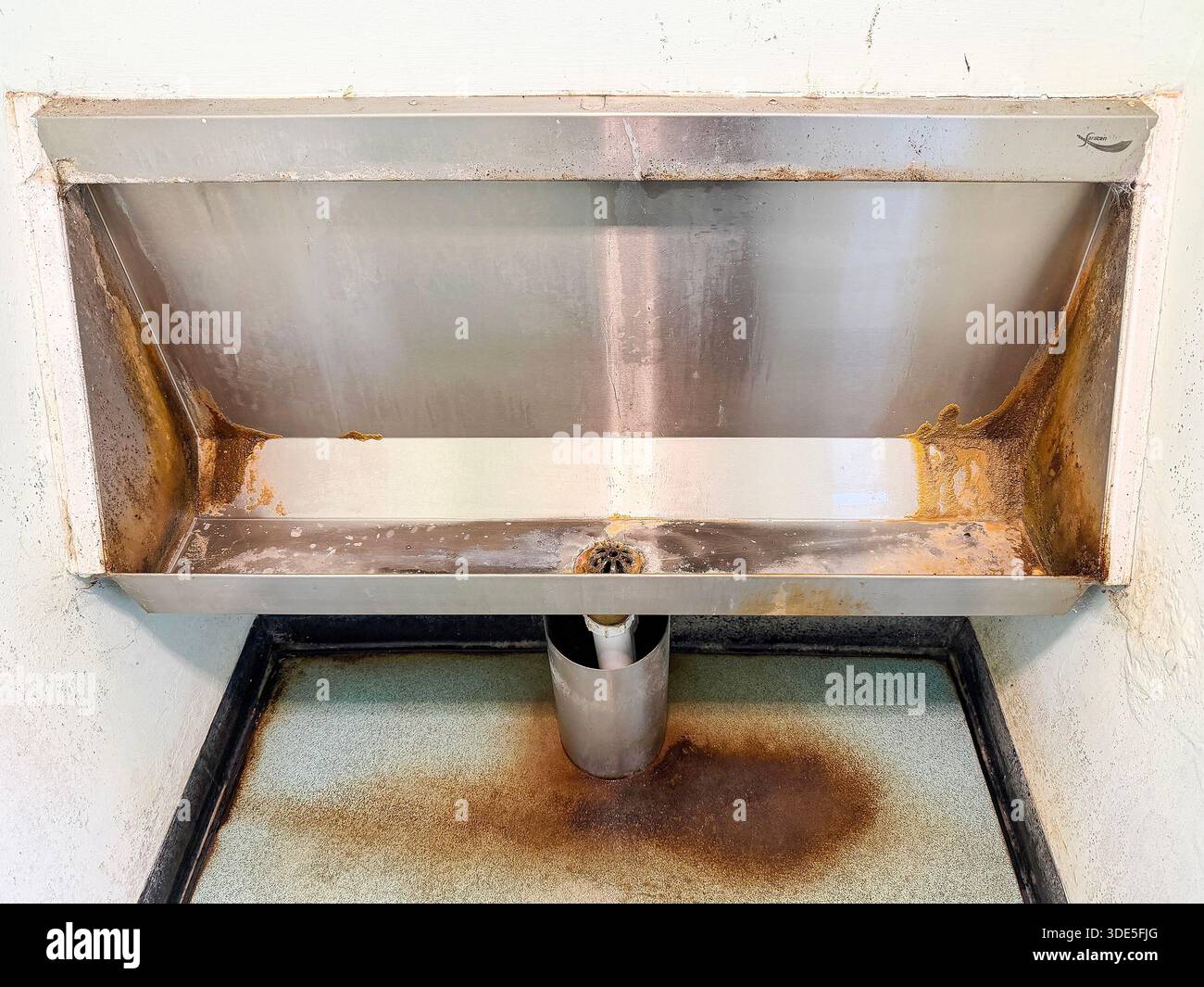 Old wall-mounted stainless steel urinal with visible rust stains and water damage in a neglected public restroom northumberland uk - Smartphone Captured Stock Image