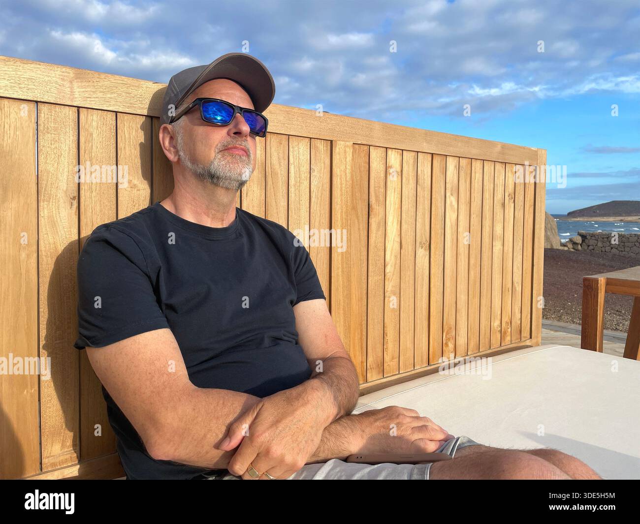 Senior older man in reflective sunglasses & cap relaxing on outdoor lounge chair next under partly cloudy sky near coastal in tenerife spain - Smartphone Captured Stock Image