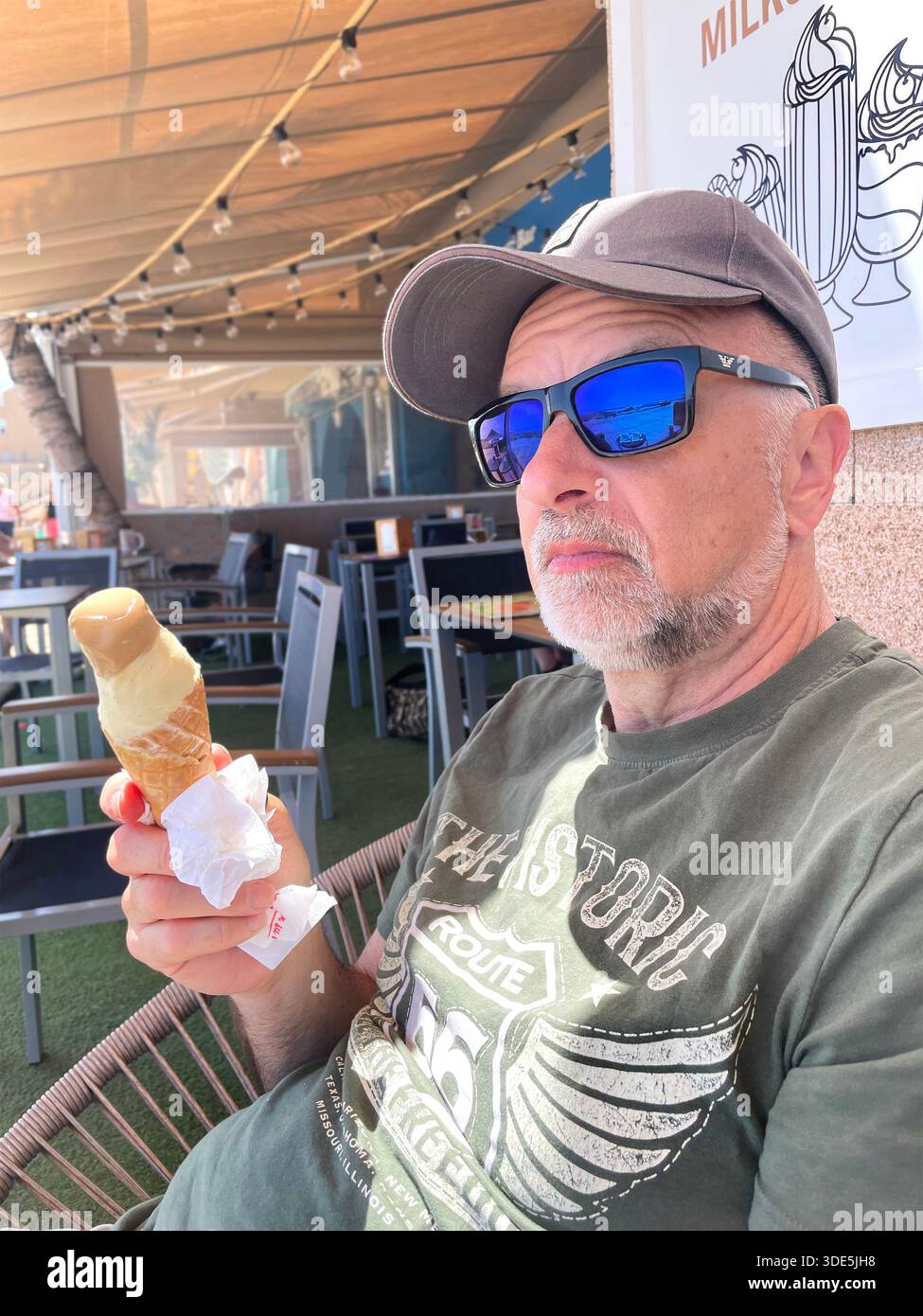 Older man wearing blue mirror sunglasses and cap enjoying ice cream cone while sitting at an outdoor café on a warm day in tenerife, spain - Smartphone Captured Stock Image