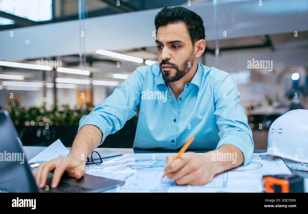Engineer Works on Project Plans in Office Setting During Daytime While Using Laptop and Pencil Stock Photo