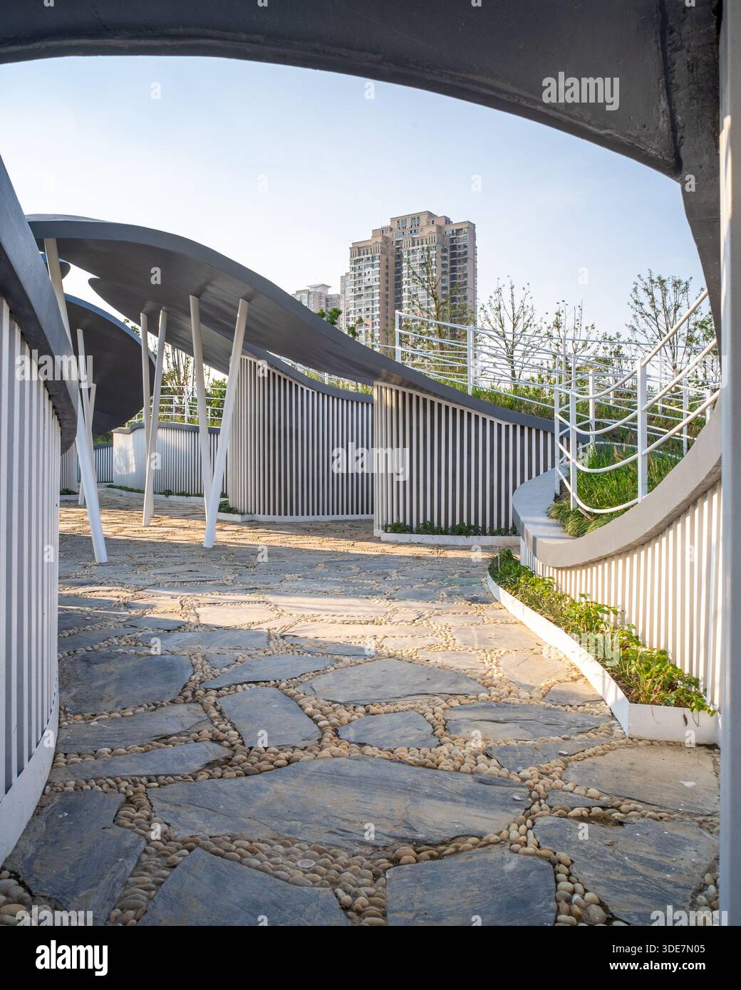 Vertical view of a stone paved path winding through a modern white corridor structure in a residential park. The image conveys urban living and commun Stock Photo