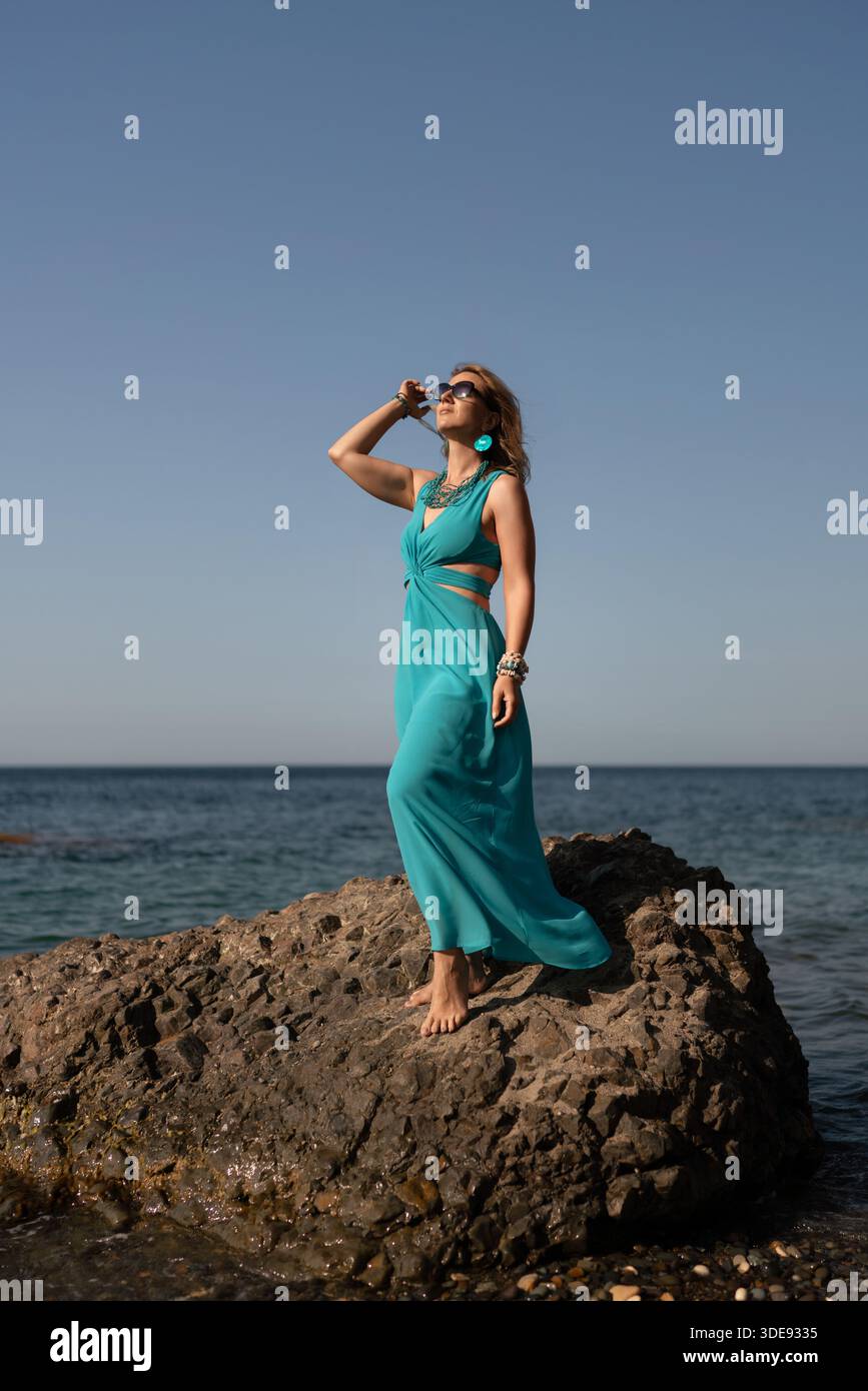 Woman, beach, vacation. Elegant woman in turquoise dress posing on rock by  the sea, enjoying summer freedom and serene ocean views Stock Photo - Alamy