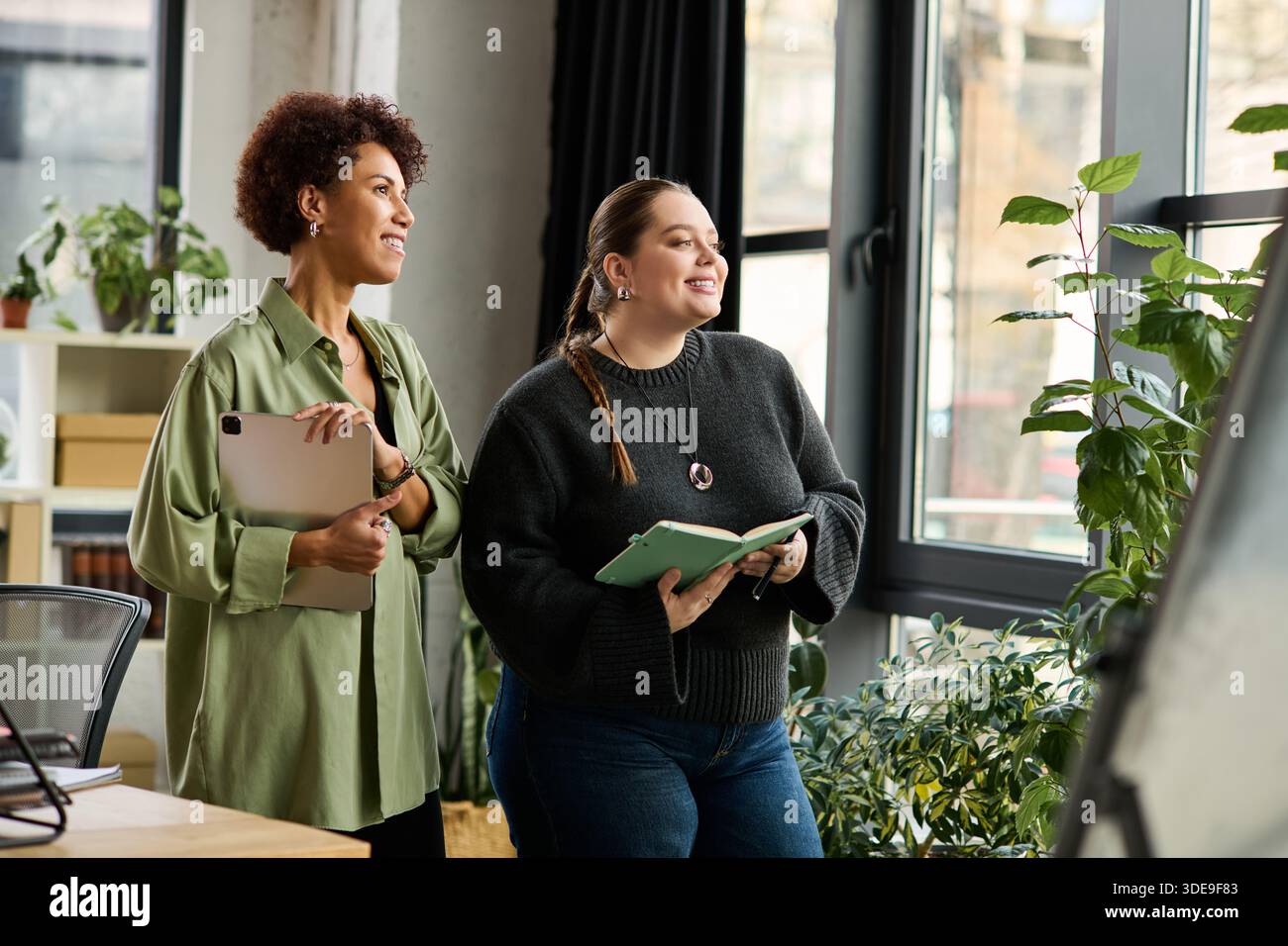 Two female managers share ideas and smiles while working in a vibrant office environment. Stock Photo