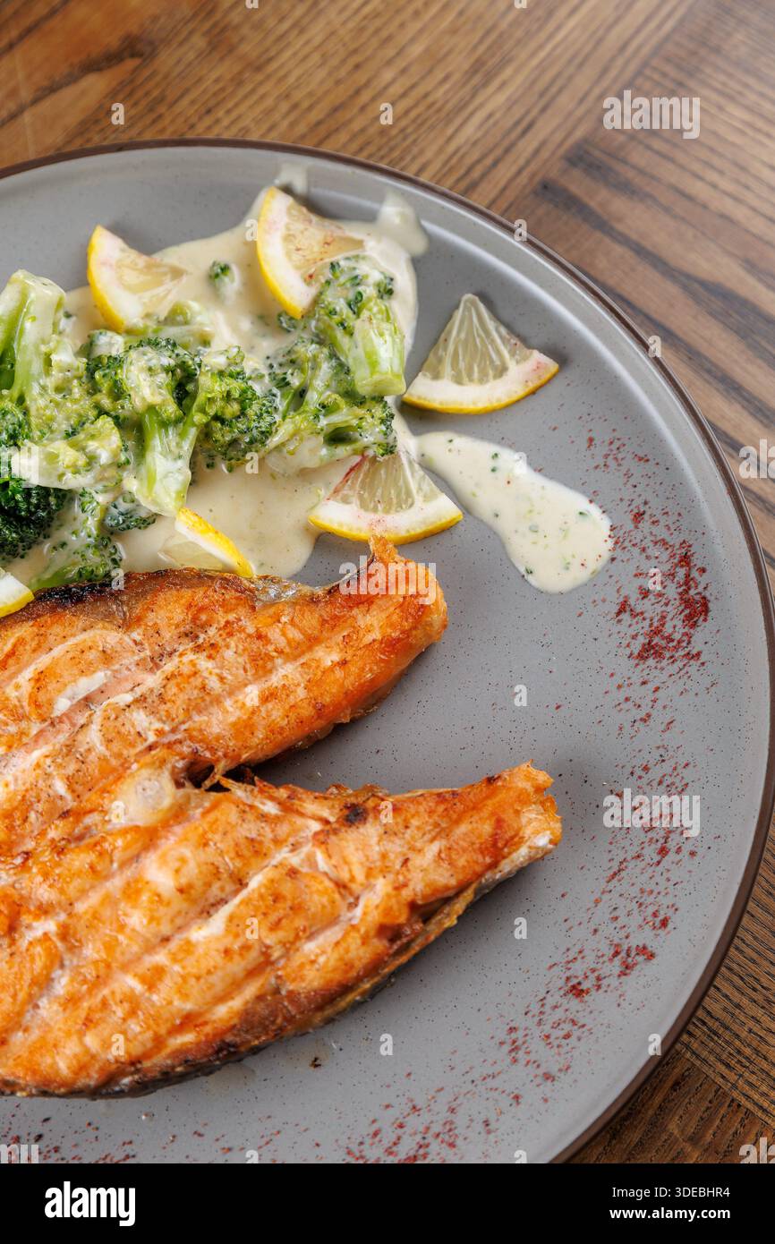 A plate features grilled fish fillets next to a creamy broccoli dish garnished with lemon slices. The setting showcases natural wood table textures. Stock Photo