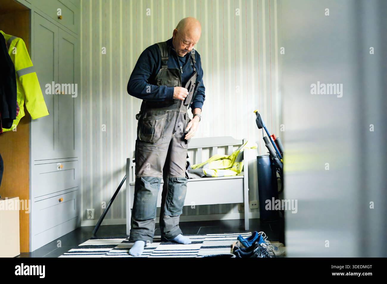Elderly male in gray overalls is getting ready in a well-lit room, with safety equipment and tools nearby, showcasing a moment of preparation and focu Stock Photo