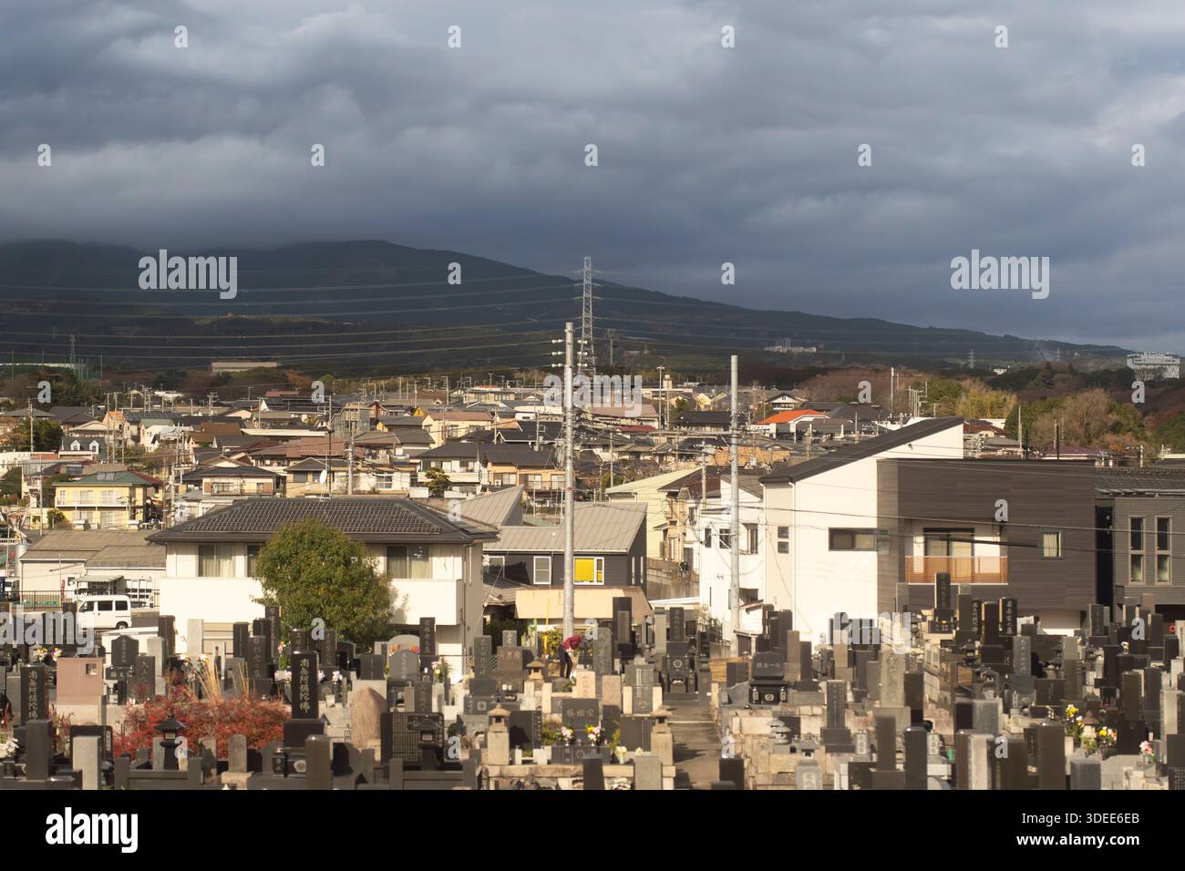 A person tends to graves in a cemetery in Mishima, Japan, with Mt. Fuji looming in the distance under a cloudy sky. Stock Photo