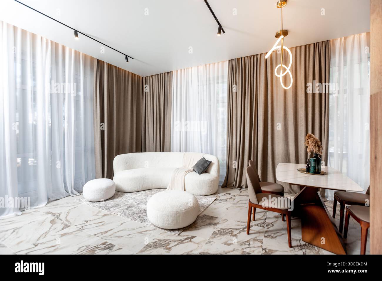A living room with a white couch, a coffee table, and a dining table. The room is decorated with white curtains and a gold lamp. The atmosphere is coz Stock Photo