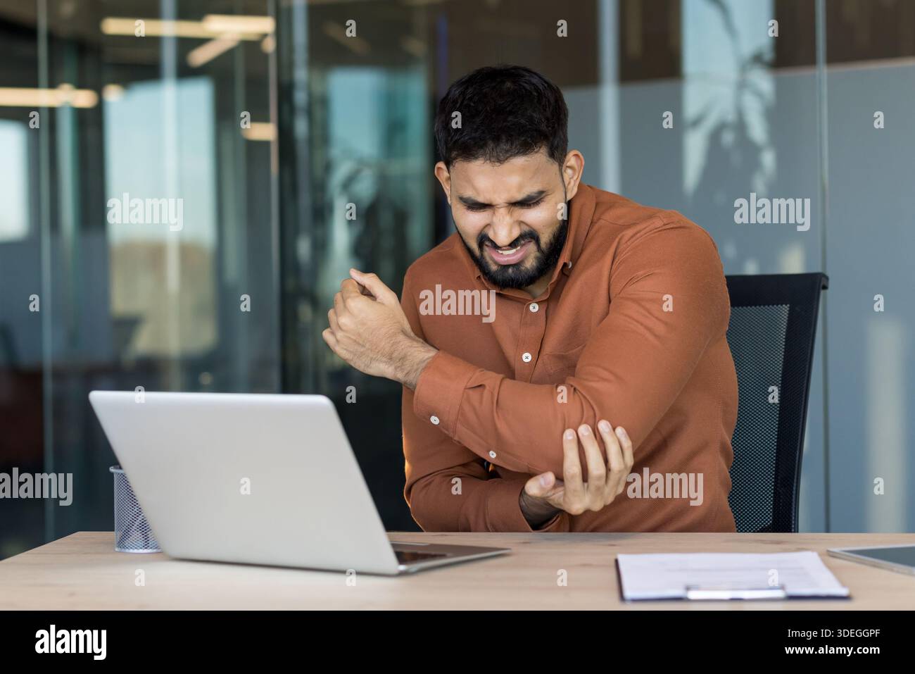 Young indian businessman clutching his elbow in pain while working on a laptop at his desk, suggesting repetitive strain, tendonitis or chronic discomfort from sedentary office work Stock Photo