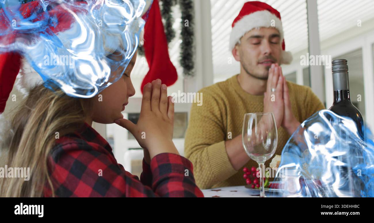 Praying dad-daughter pressing hands at home table, wearing Santa hats, mustard sweater, wine bottle Stock Photo