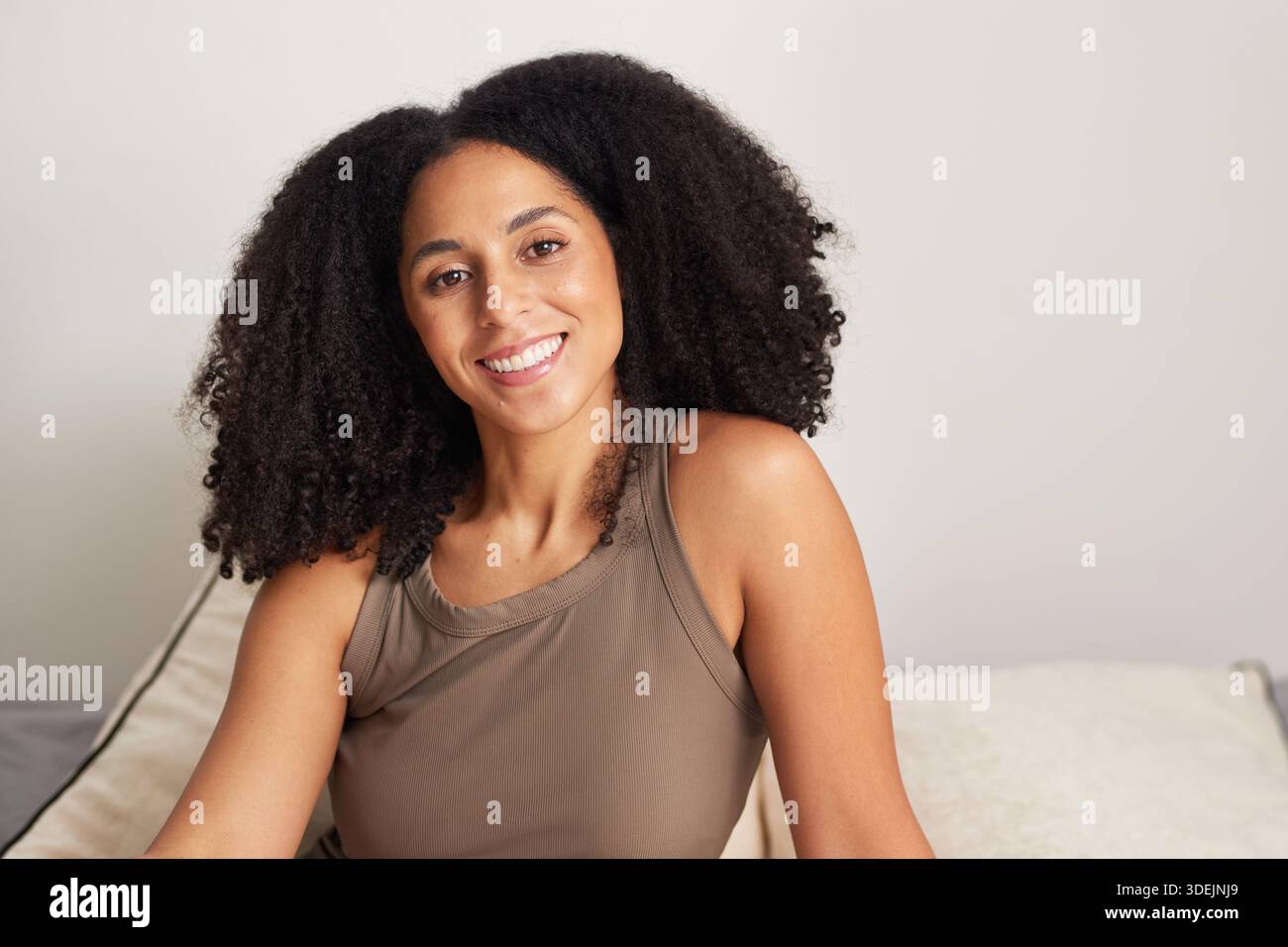 Smiling Woman With Curly Hair In Brown Tank Top Sitting On Couch At Home Stock Photo