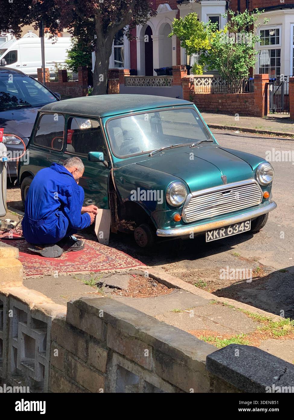 Mechanic in blue overalls repairing a vintage green mini cooper on a residential street in England the front wheels removed working on knees and rug - Smartphone Captured Stock Image