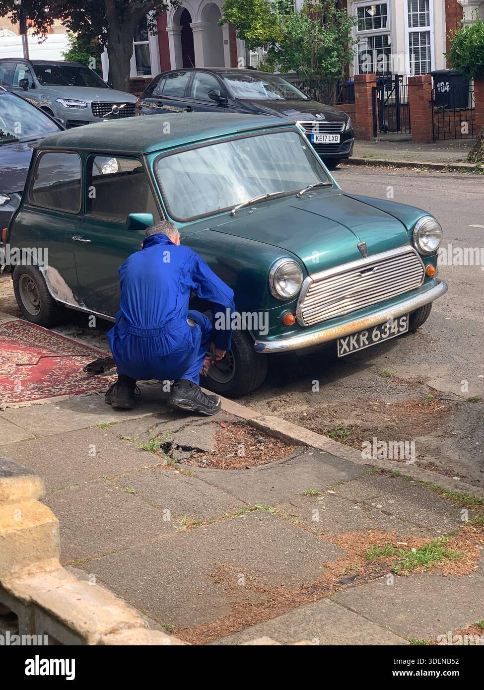 Mechanic in blue overalls repairing a vintage green mini cooper on a residential street in England the front wheels removed working on knees and rug - Smartphone Captured Stock Image