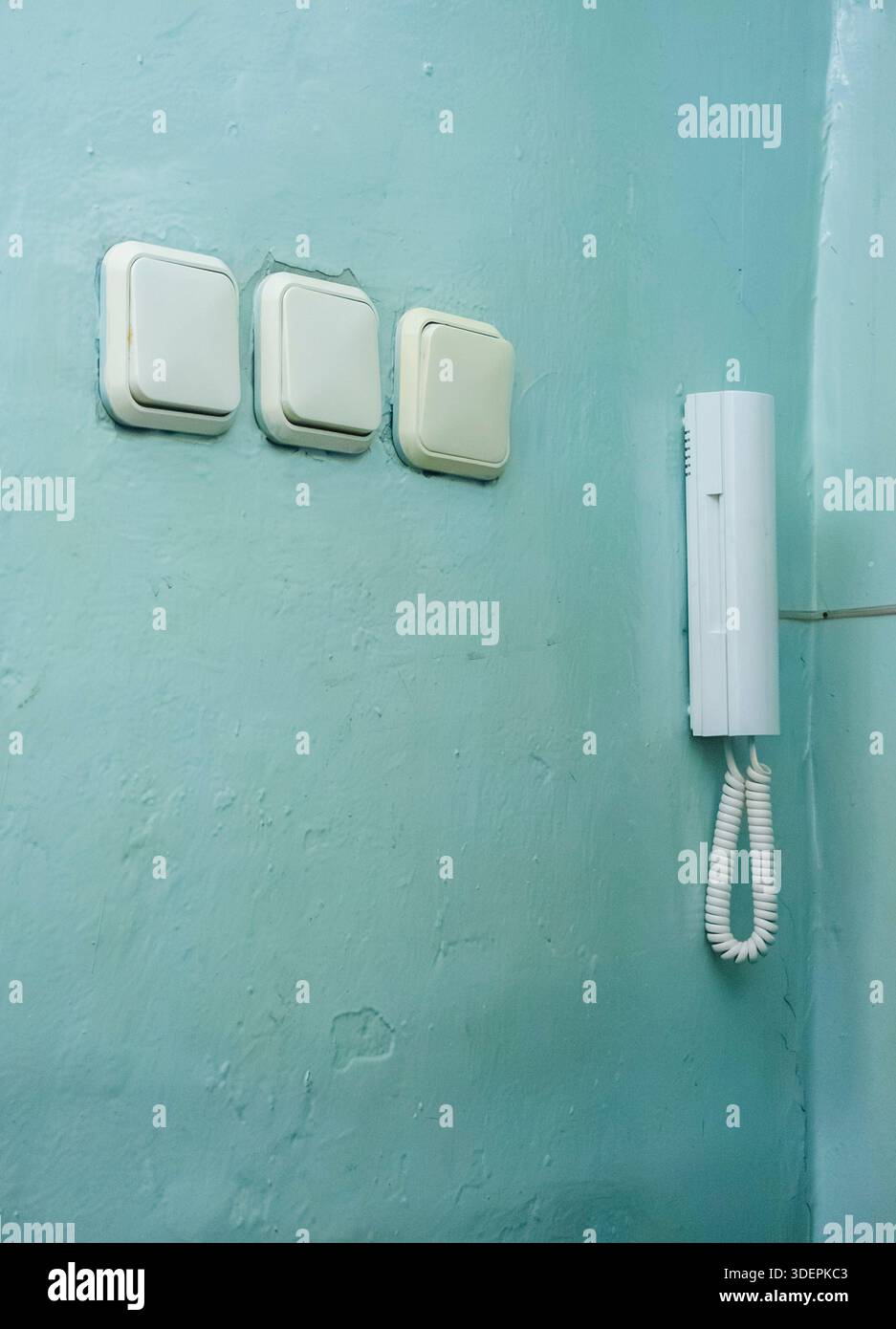 Three light switches and an old white wired intercom are mounted on a worn, textured aqua colored wall, showing everyday functional elements in a resi Stock Photo