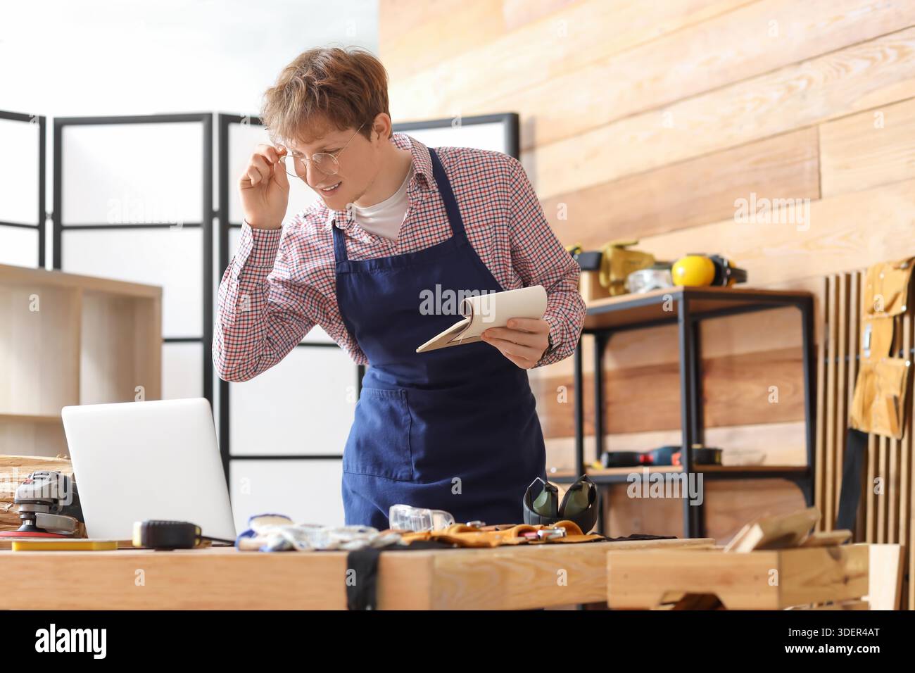Young carpenter with notebook using laptop at table in workshop Stock Photo