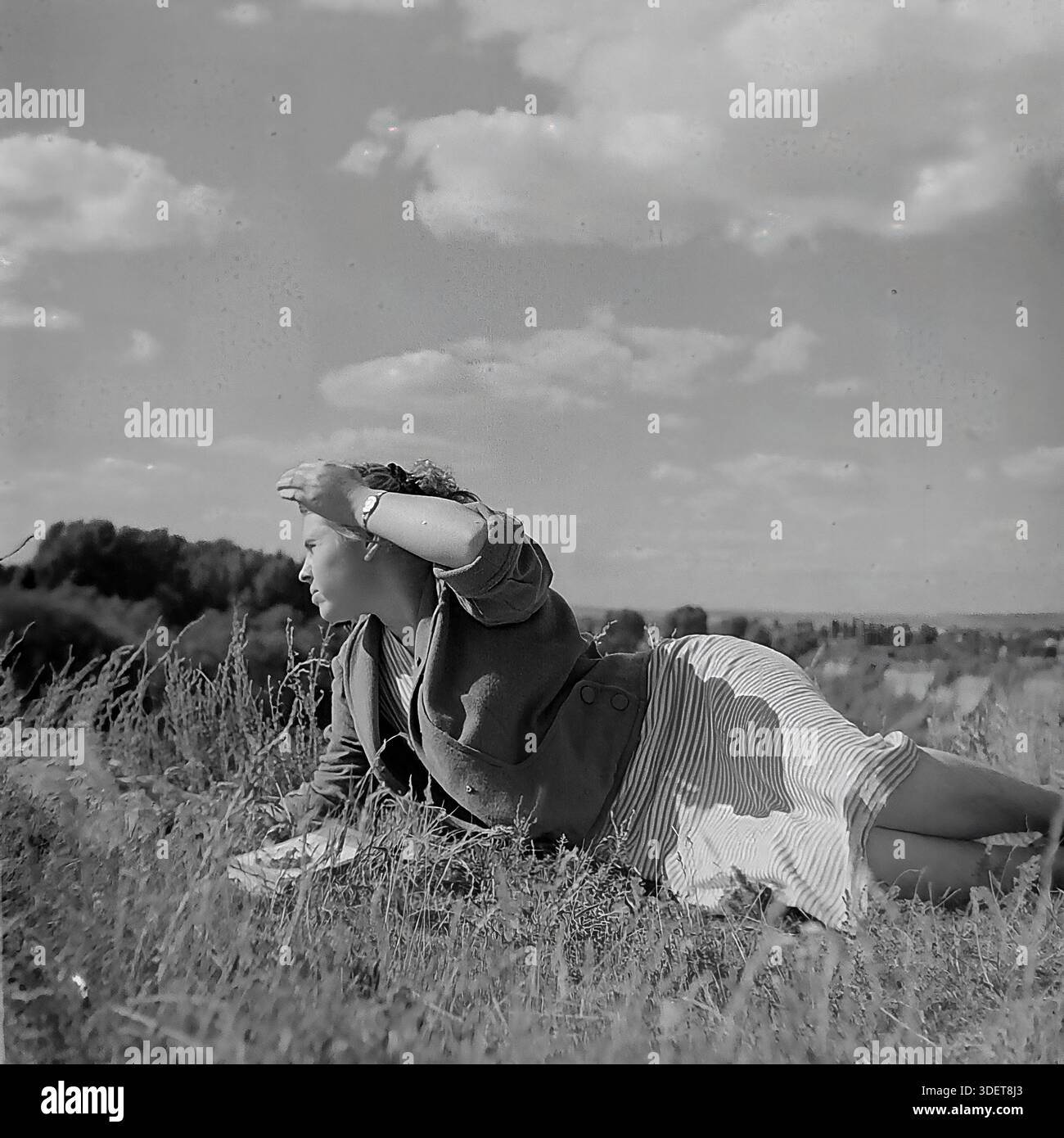 Slaviansk, Donetsk region, Ukraine, USSR. 1950s. Artistic archival photo of Valentina Timofeevna Kravelis resting on the high bank of the Kazeny Torets river. The young woman lies on the grass in a stylish striped dress, shielding her eyes from the bright summer sun with her hand. The vast landscape of the river valley opens up in the background. The image captures the aesthetics of Soviet fashion, the carefree mood of youth and the serene atmosphere of the peaceful Donbas during the stable period of life before the war. Stock Photo