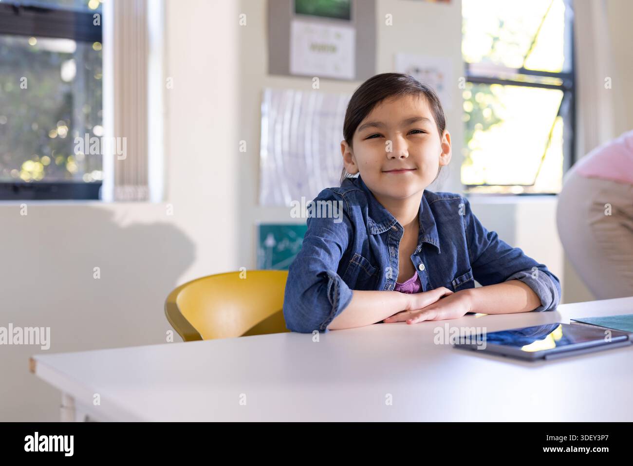Pre-teen female student sitting on yellow chair at white class table in denim jacket, tablet nearby Stock Photo