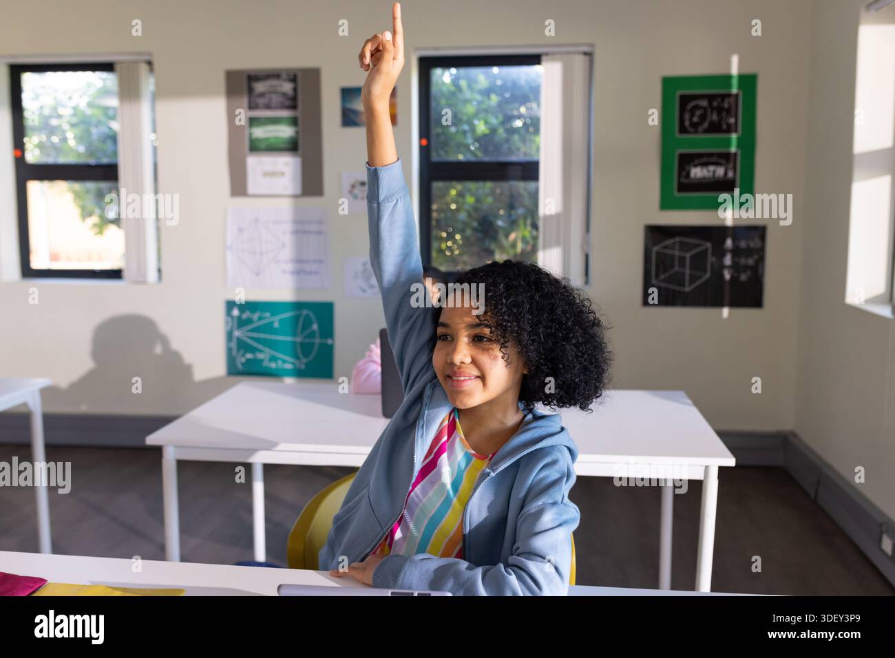 African American child in blue hoodie raising right hand sitting in classroom with math posters Stock Photo