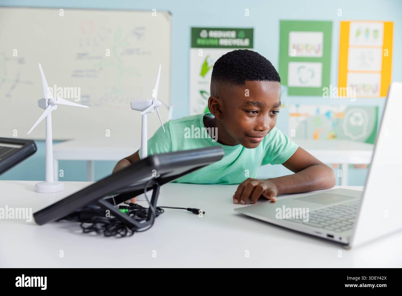 African male youth testing laptop, wind turbines and black charger in STEM lab wearing green tee Stock Photo