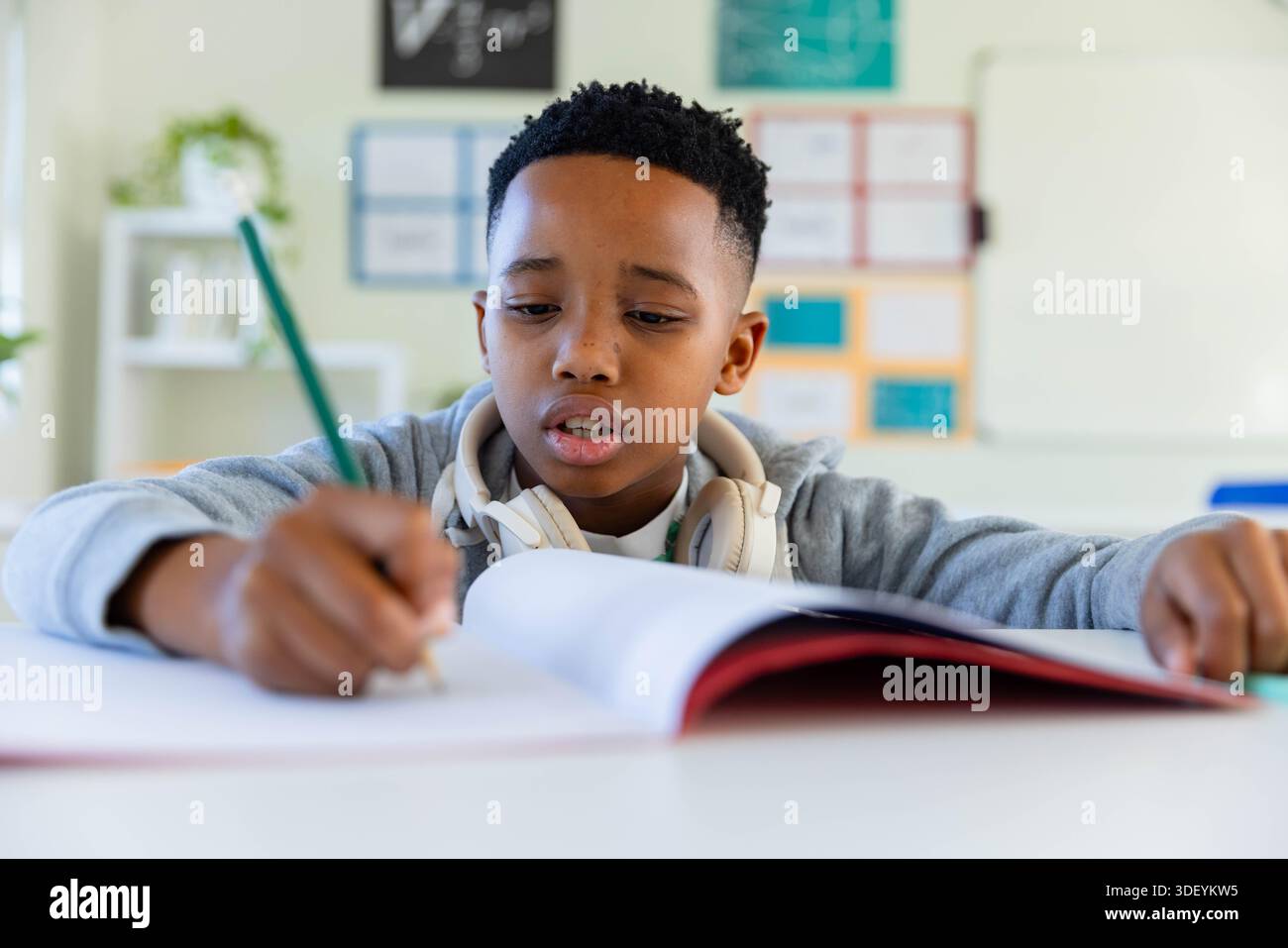 Preteen African American boy writing in workbook at low desk with green pencil and headphones Stock Photo