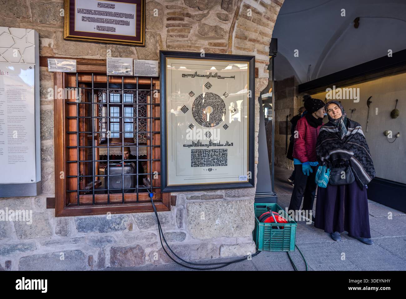 The Mevlana Museum (Mevlânâ Müzesi) in Konya's Karatay district, Turkey, houses a traditional dervish room with a barred window. A young man sits behind it, reciting verses from the Koran that carry across the museum courtyard via loudspeaker. The museum is the mausoleum of the Sufi mystic Rumi (Jalāl ad-Dīn ar-Rūmī, 1207–1273), with an adjoining mosque and former dervish lodge; the turquoise dome built over his grave in 1274 is considered a symbol of Konya. Stock Photo
