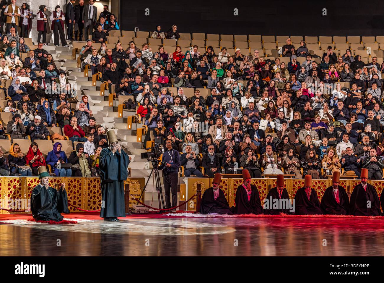 The end of the Şeb-i Arus ceremony at the Mevlana Kültür Merkezi in Karatay, Konya, Turkey, a venue that holds up to 3,000 people, is heralded by a recitation from the Koran. After their whirling dance, the dervishes are once again cloaked in their black robes. A dervish kneels with his hands folded before his face on the red carpet, while a master of ceremony stands beside him and the applauding audience is visible in the background. Şeb-i Arus is the annual festival marking the death anniversary (17 December) of the mystic poet Rumi (Jalāl ad-Dīn ar-Rūmī, 1207–1273), founder of the Mevlevi o Stock Photo