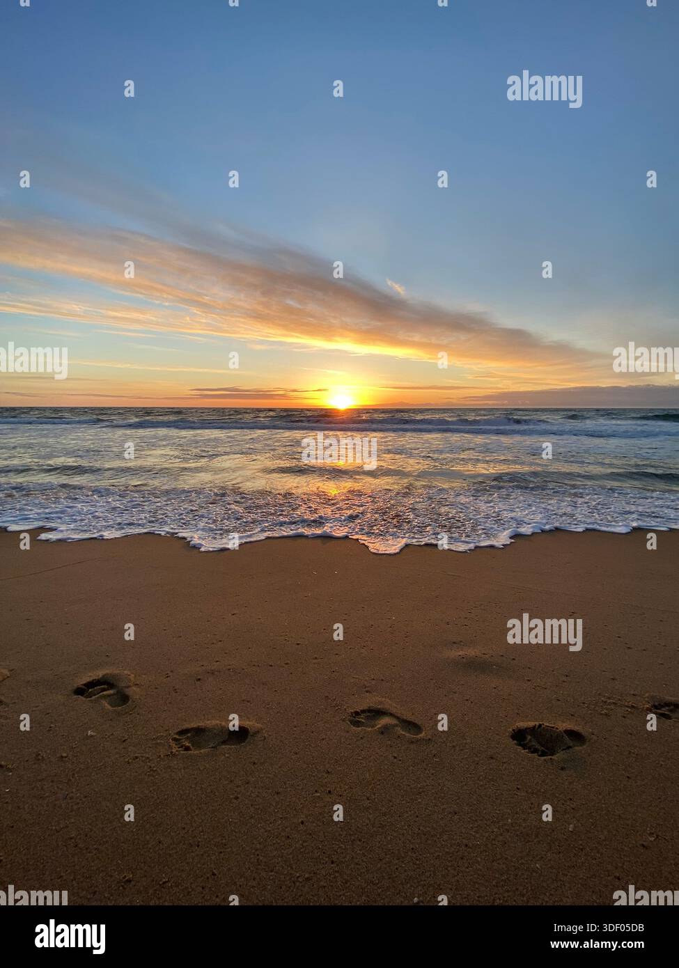 Footprints in sandy beach leading toward ocean waves at sunrise, with golden light reflecting on the water. - Smartphone Captured Stock Image