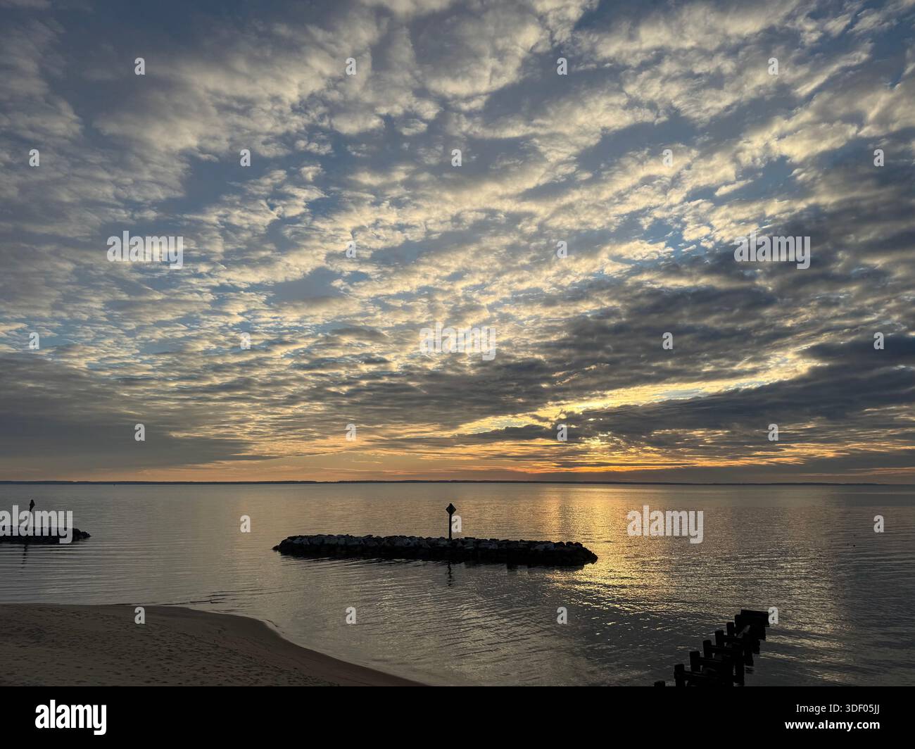 Golden sunrise over a calm bay with dramatic textured clouds and stone breakwater silhouettes reflecting on tranquil water. - Smartphone Captured Stock Image