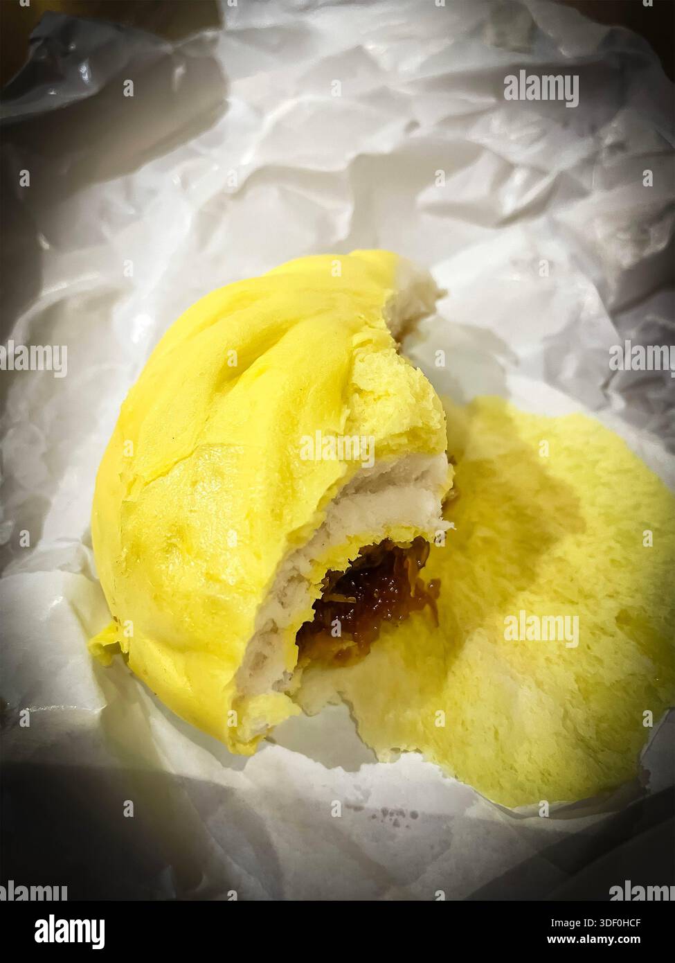 Partially eaten steamed yellow bao  bun with sweet or savory filling placed on crumpled white paper, malaysia - Smartphone Captured Stock Image