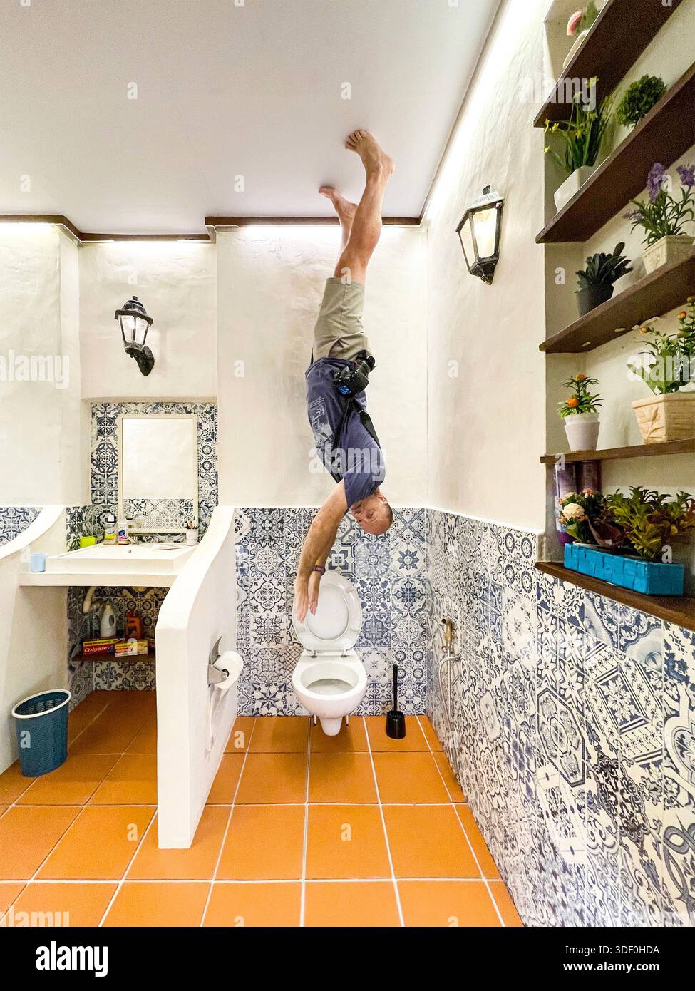 Man appearing to stand on his hands on the ceiling of an upside-down themed bathroom with patterned tiles and decor - Smartphone Captured Stock Image