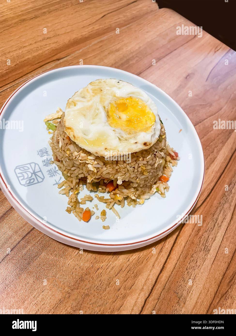 Plate of nasi goreng, which is Indonesian Malaysian-style fried rice with mixed vegetables and fried egg on top served on wooden table in a restaurant - Smartphone Captured Stock Image