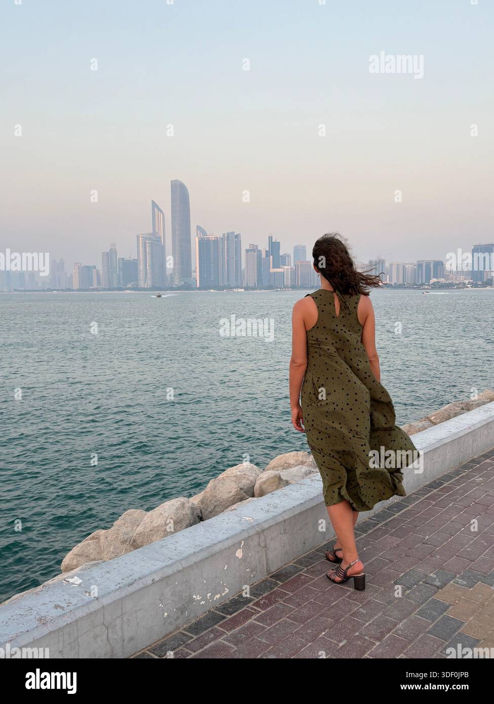Woman in olive green gazes across the waterfront toward Abu Dhabi’s shimmering towers at dusk - Smartphone Captured Stock Image