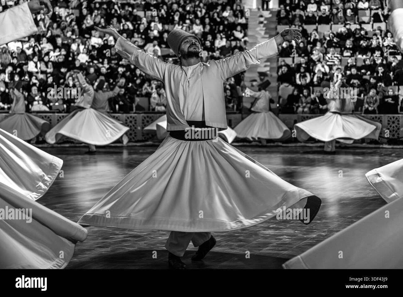 The Şeb-i Arus ceremony at the Mevlana Kültür Merkezi in Karatay, Konya, Turkey, a venue that holds up to 3,000 people, shows a dervish with raised arms and his gaze turned fully upward in the foreground during the ritual Sema dance, his skirt swinging wide in full rotation. The right hand points upwards, receiving God's blessing. The left hand points downwards, passing it on to the earth. The dervishes spin counter-clockwise, symbolically against the ego. The right foot stays fixed while the left foot drives the rotation. Şeb-i Arus is the annual festival marking the death anniversary (17 Dec Stock Photo