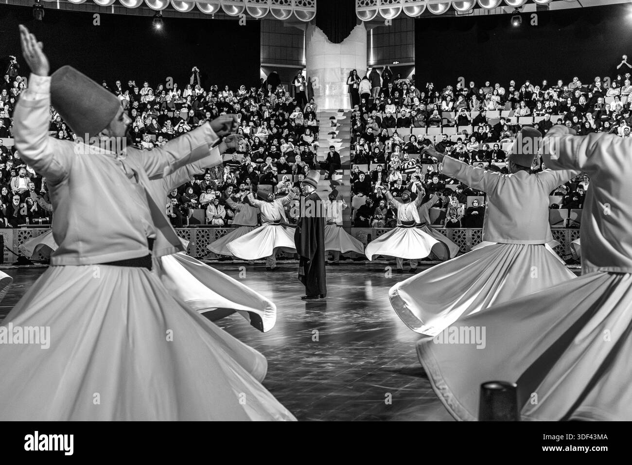 The Şeb-i Arus ceremony at the Mevlana Kültür Merkezi in Karatay, Konya, Turkey, a venue that holds up to 3,000 people, shows the Gözcü (overseer) remaining amid the dancers in a dark robe, framed by the raised arms of several dervishes during the ritual Sema dance. The right hand points upwards, receiving God's blessing. The left hand points downwards, passing it on to the earth. The dervishes spin counter-clockwise, symbolically against the ego. The right foot stays fixed while the left foot drives the rotation. Among the dancers, the Gözcü usually remains inconspicuous to the audience; he p Stock Photo