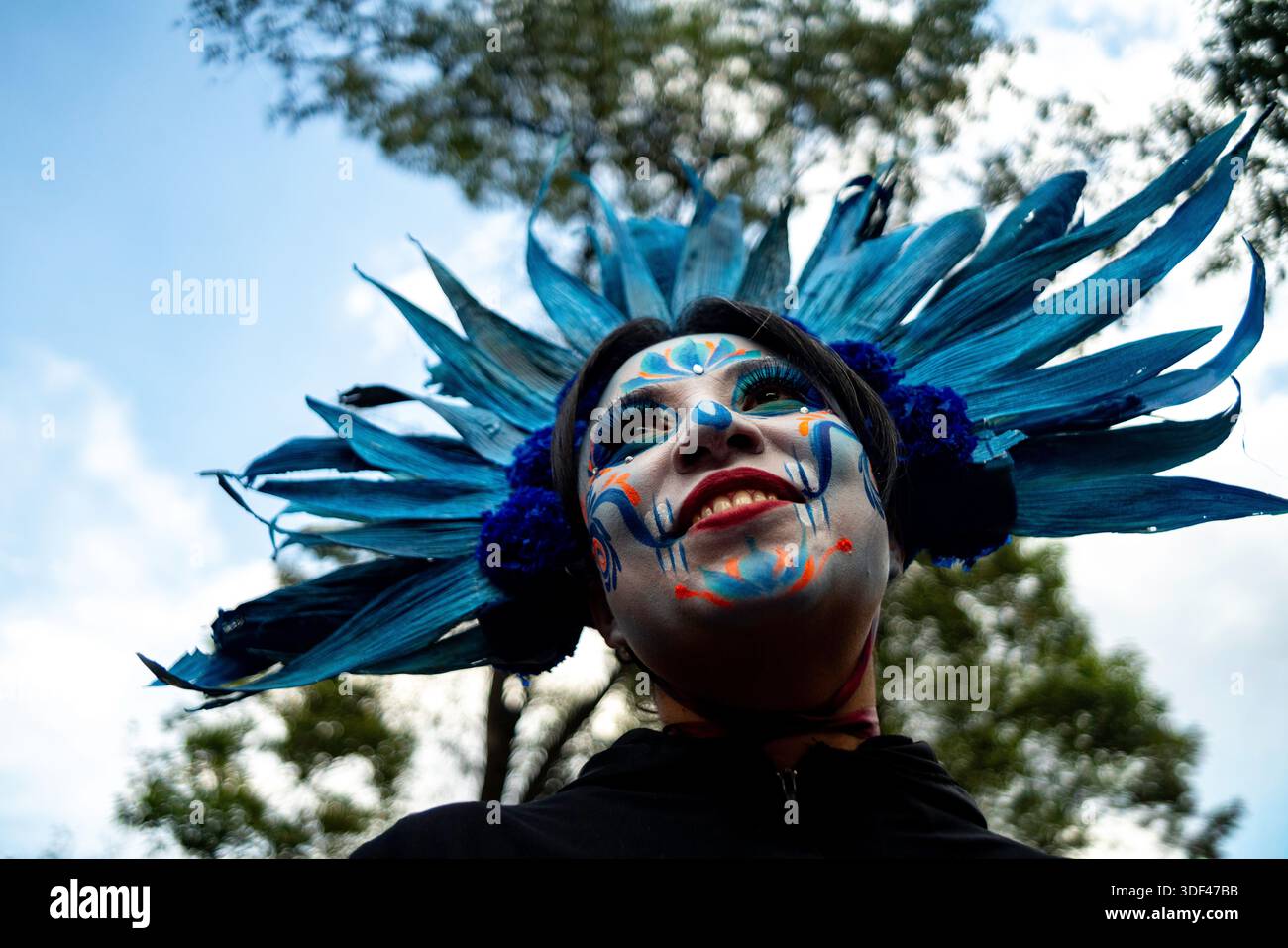 Low angle portrait of a participant wearing skull makeup and a blue headdress during the Day of the Dead parade in Mexico City, Mexico. Stock Photo